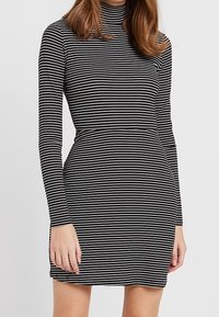 Mustavalkoraidallinen mekko, jossa on pitkät hihnat ja korkea kaulus. Istuva siluetti, jossa on hieman levenevä hame ja pehmeä rakenne.