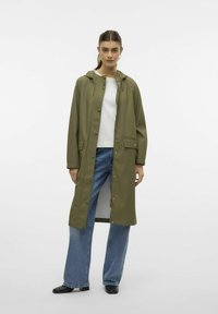 Olivno zeleni vodoodporni plašč s kapuco, pritisnimi gumbi, velikimi sprednjimi žepi in ravno silhueto, v kombinaciji s svetlo modrimi kavbojkami.