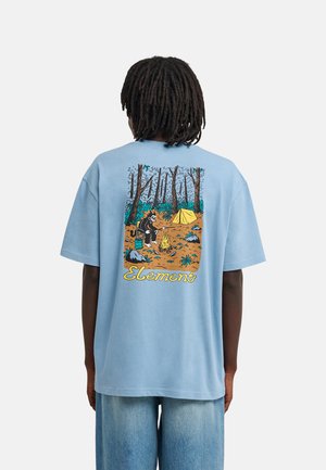 Personne avec des dreadlocks portant une chemise bleu clair avec une scène de camping en forêt et le mot "Element" imprimé dans le dos.