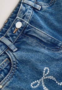 Shorts di denim blu medio con finitura testurizzata. Presentano un bottone argentato, passanti per cintura e dettagli decorativi con strass.