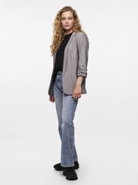 Vrouw met krullend haar draagt een grijze blazer, een zwart shirt, lichtblauwe spijkerbroek en zwarte schoenen, staand tegen een witte achtergrond.