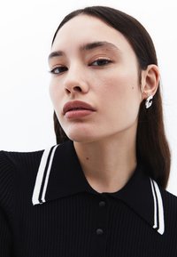 Svart ribbad polo med vitrandig krage. Modellen bär en silverörhänge i form av en stiliserad våg. Slät hy och neutral sminkning.