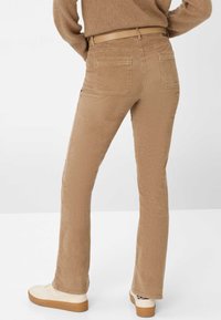 Person iført beige kordfløjl bukser, tan bælte, brun sweater og hvide sneakers, stående med hænderne på hofterne foran en hvid baggrund.