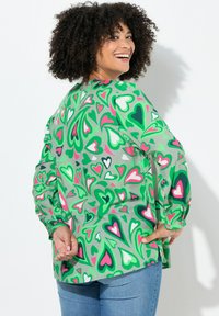 Grüne Bluse mit Herzmuster in Pink- und Weißtönen. Hat lange Ärmel und eine lässige Passform. Das Material wirkt leicht.