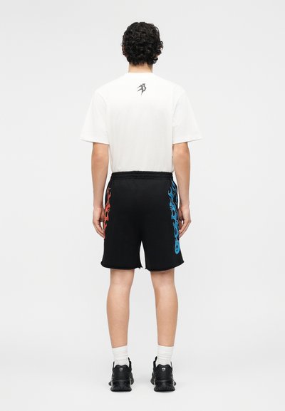 Άτομο με σγουρά σκουρόχρωμα μαλλιά στέκεται γυρισμένο προς τα πίσω φορώντας άσπρο μπλουζάκι και μαύρο σορτς με κόκκινα και μπλε σχέδια φλόγας στα πλάγια.