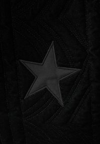 Tejido negro con un diseño acolchado y sutiles patrones en forma de ola. Una prominente estrella negra está cosida en el centro, mostrando una textura suave.