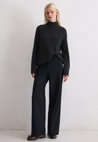 Pull en maille côtelée noir avec col montant, associé à un pantalon noir à jambes larges. Texture douce, coupe décontractée et design minimaliste. Chaussures brunes incluses.