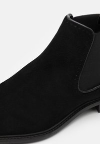 Bottine en suède noire avec des panneaux latéraux élastiques, une texture lisse, un bout pointu et un talon étroit empilé. Détails de couture subtils visibles.
