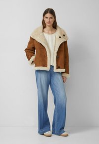 Brązowa zamszowa kurtka z kożuchem i puszystym kremowym kołnierzem, zapinana na dwa duże guziki, noszona na lekkim swetrze i zestawiona z szerokimi niebieskimi dżinsami.