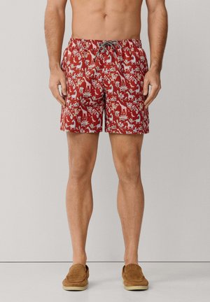 Man draagt rood korte broek met dieren- en plantenprint, staat op blote voeten tegen een lichte achtergrond.