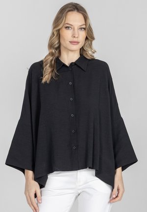 Kobieta o długich blond włosach nosi luźną czarną bluzkę zapinaną na guziki z szerokimi rękawami oraz białe spodnie, stojąc na szarym tle.