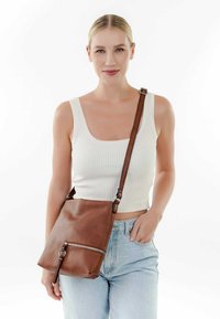 Sac en cuir marron à bandoulière avec une finition texturée, comportant une poche zippée à l'avant et une sangle ajustable, présenté sur fond blanc.