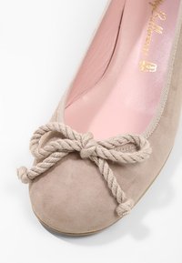 Ballerine en daim beige avec un gros nœud en corde sur l'orteil et une semelle intérieure rose rembourrée avec un logo doré.
