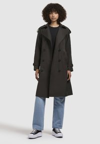 Trench coat de culoare verde închis, cu design double-breasted, butoane mari, epauleți pe umeri și buzunare laterale, purtat cu blugi albastri deschis și adidași negri.
