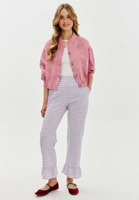 Różowa kurtka zapinana na guziki z dekoracyjnymi wycięciami, biała koszula pod spodem, lawendowe spodnie w kratkę z falbanami na dole i czerwone baleriny.