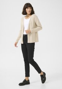 Beige langetermijn cardigan van zacht materiaal, open voorkant ontwerp, gedragen over een witte top en gecombineerd met zwarte skinny jeans en zwarte schoenen.