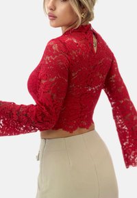 Cropped top in pizzo rosso con maniche lunghe a campana, dotato di uno schienale a chiave e un motivo floreale, abbinato a pantaloni aderenti di colore chiaro.