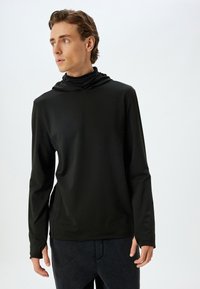 Camisa atlética negra de manga larga con capucha, que presenta una textura suave y un diseño ajustado, combinada con pantalones oscuros.