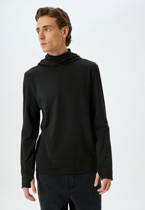 Camisa atlética negra de manga larga con capucha, que presenta una textura suave y un diseño ajustado, combinada con pantalones oscuros.