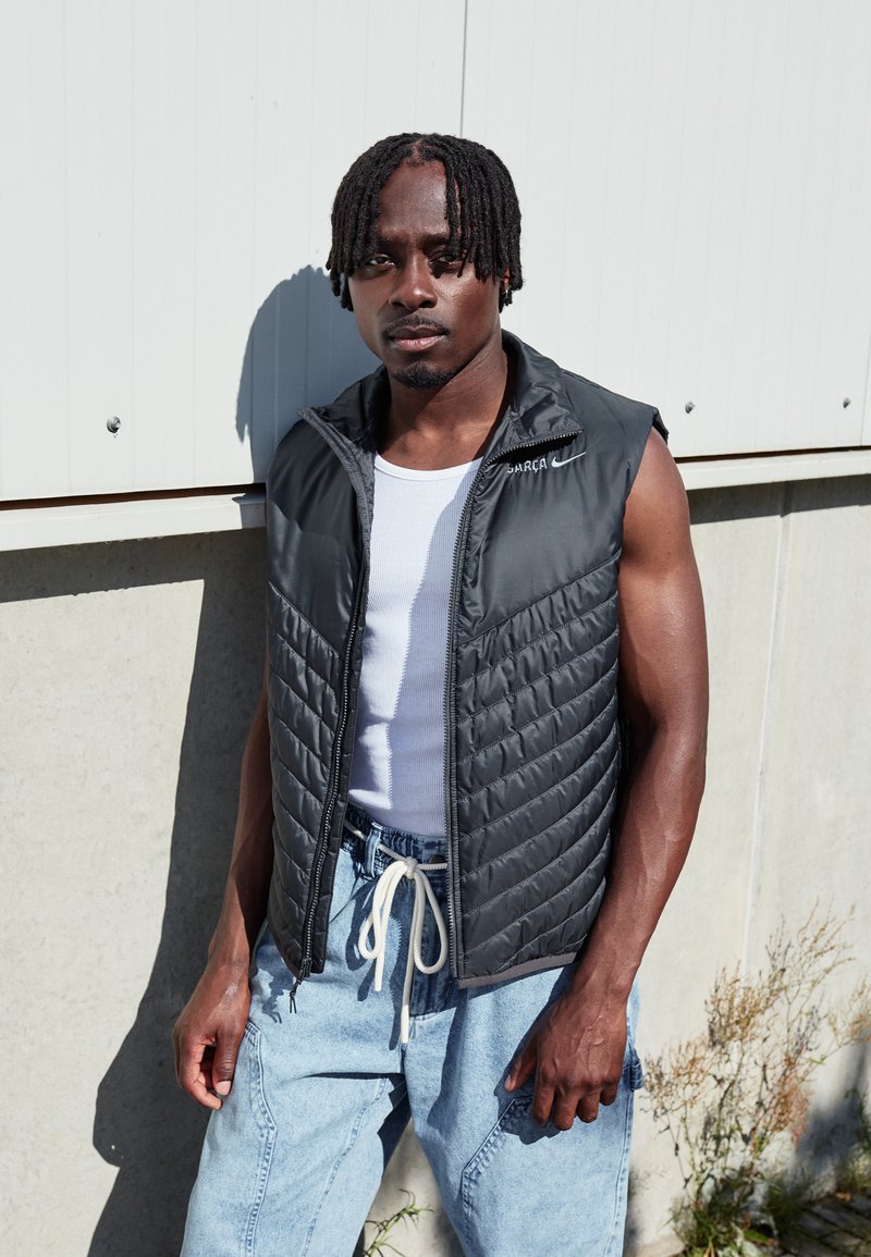 Black quilted sleeveless vest with a high collar and zip closure, layered over a white tank top, paired with blue denim pants.