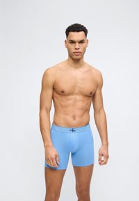 Άνδρας που στέκεται μπροστά σε απλό φόντο, φορώντας ανοιχτό μπλε μποξεράκια Calvin Klein μέχρι το μέσο του μηρού, κοιτάζοντας μπροστά με ουδέτερη έκφραση.