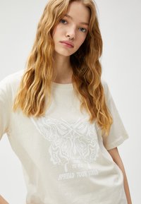 Cremefarvet bomulds t-shirt med et stort hvidt sommerfugle-tryk og tekst nedenunder, der står "Du er fri til at flyve. Sprede dine vinger."