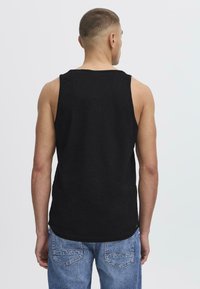 Musta tank top, joka on valmistettu puuvillasta, jossa on pyöreä pääntie ja hihaton muotoilu. Takana on pyöristetty helma ja minimaalinen yksityiskohtaus.