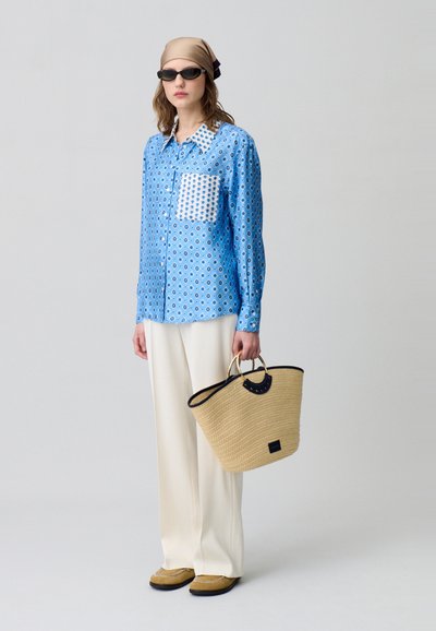 Camicia a maniche lunghe blu con motivo, taschino bianco, pantaloni larghi bianchi, borsa di paglia con bordi neri e scarpe in suede marroni. Occhiali da sole e foulard.