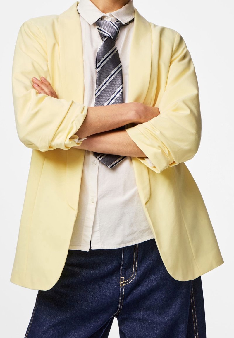 Personne les bras croisés portant un blazer jaune clair, une chemise blanche boutonnée, une cravate rayée et un jean bleu foncé sur fond blanc.