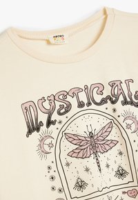 Sudadera infantil color crema con un gráfico de libélula, estrellas y diseños de luna en impresión negra y rosa. Detalle de cuello acanalado.