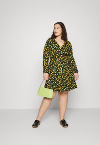 Vestido preto com padrão de fruta amarela e verde, mangas longas e design envolvente; combinado com uma mala verde-clara e tamancos pretos.