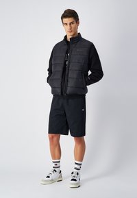Schwarze Steppweste mit gestepptem Design, Reißverschluss vorne, kombiniert mit einem schwarzen Sweatshirt, schwarzen Shorts, weißen Sneakern und gestreiften Socken.
