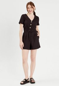 Jumpsuit short noir avec un décolleté en V, des manches courtes, quatre boutons sur le devant, une taille ceinturée et des poches latérales. Porté avec des sandales noires.
