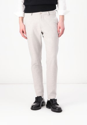 Mand iført lysegrå slim-fit bukser, sorte klaksko, hvid skjorte og sort ærmeløs sweater, stående mod en hvid baggrund.