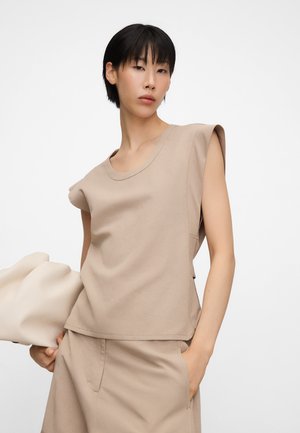 Femme aux cheveux noirs courts portant un haut beige sans manches et un short assorti, tenant une grande pochette beige sur fond blanc.