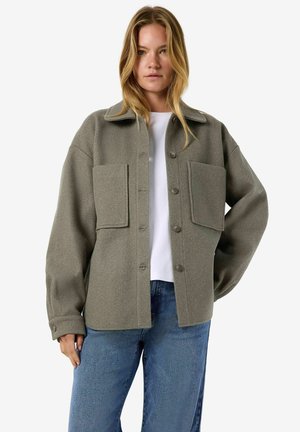 Femme debout, une main sur la hanche, portant une veste boutonnée vert olive avec de grandes poches devant, une chemise blanche et un jean bleu.