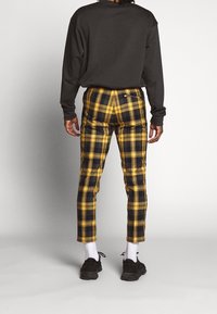 Camisa negra de manga larga, pantalones de cuadros amarillos y negros con ajuste entallado, con un bolsillo trasero, combinados con calcetines blancos y zapatillas negras.