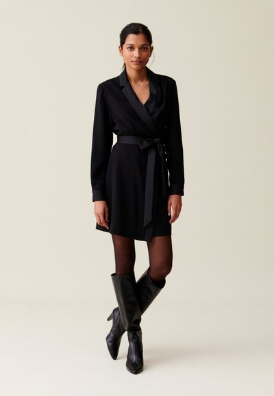 Abito avvolgente nero con maniche lunghe, colletto in satin e cinture coordinate. Stivali neri lucidi al ginocchio completano il look, sullo sfondo neutro.