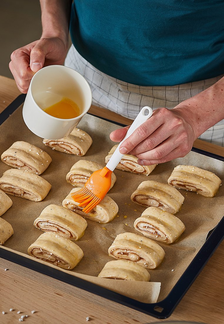 Backblech mit ausgerollten Teigstücken, die mit Zimtfüllung belegt sind. Eine Hand gießt flüssiges Ei in eine Tasse und benutzt einen orangefarbenen Backpinsel.