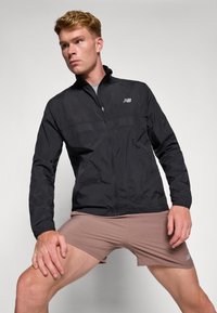 Sort letvægtsjakke med lynlås foran, med et mesh-panel over brystet og et logo på venstre side, båret over beige shorts.