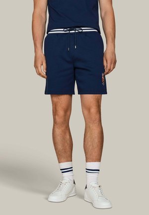 Hombre con pantalones cortos azul marino con cordón, calcetines blancos con rayas azules y zapatillas blancas, de pie frente a un fondo liso.