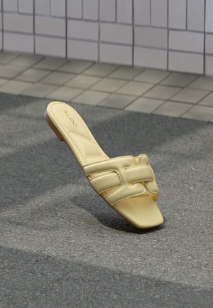 Lys gul flad sandal med brede polstrede stropper og firkantet tå, mærket "ALDO," placeret på asfalt nær flisebelagt fortov.
