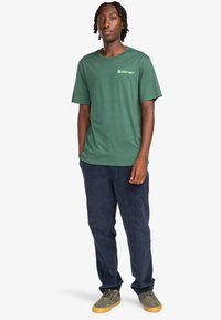 Camiseta de algodón verde con cuello redondo y mangas cortas, que presenta un pequeño logo en verde claro. Combinada con pantalones de pana en color azul marino y zapatos verdes.