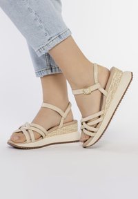 Beige wedge sandals feature a woven jute accent, adjustable ankle strap, and multiple crisscrossing straps over the toes with a rubber sole.