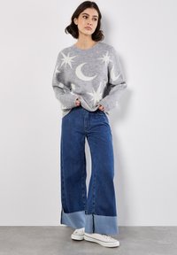 Hellgrauer Strickpullover mit Stern- und Mondmustern, kombiniert mit weit geschnittenen blauen Jeans aus Denim mit einem kontrastierenden Saum. Weiße Sneakers inklusive.