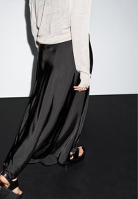 Persoon in een crème trui en een vloeiende zwarte satijnen rok die over een donkere vloer loopt, met zwarte plateauzandalen aan.