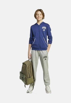Adolescent portant un sweat à capuche zippé bleu Adidas et un pantalon de jogging gris Adidas, tenant un sac à dos vert olive, se tenant debout et souriant.