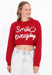 Roter, kurzer Pullover aus Strickstoff mit dem Text "Lächle jeden Tag" in Weiß, weiten Ärmeln und einem gerippten Ausschnitt.