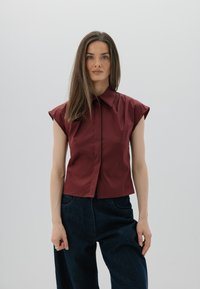 Burgundowa koszula z krótkim rękawem i spiczastym kołnierzykiem, zapinana na guziki z przodu, o gładkiej fakturze, noszona z ciemnoniebieskimi spodniami dżinsowymi z wysokim stanem.