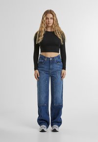 Schwarzes langärmeliges Cropped-Top, blaue weit geschnittene Jeans mit kontrastierendem Paneeldesign, silberne Hardware und graue Sportschuhe. Einfarbiger Hintergrund.